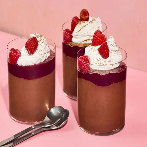 Three elegant chocolate mousse dessert cups layered with raspberry jelly, topped with whipped cream, fresh raspberries, and a dusting of cocoa, presented on a soft pink background.