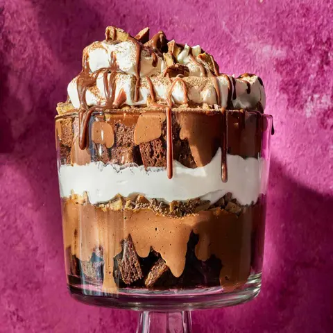 Decadent layered chocolate dessert parfait topped with toasted marshmallows, brownie chunks, and melted chocolate drizzle in a glass cup, set against a vibrant pink textured background.